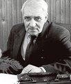 Man with white hair and a dark suit on next to a typewriter looking at the camera.