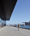 A view of the Opera House from a distance. In front of the opera house, there is the harbour. There are people walking around. It looks like summer.