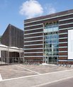 A photo of a big square brown brick building. In the middle are a row of windows. In front of the building are many bikes.